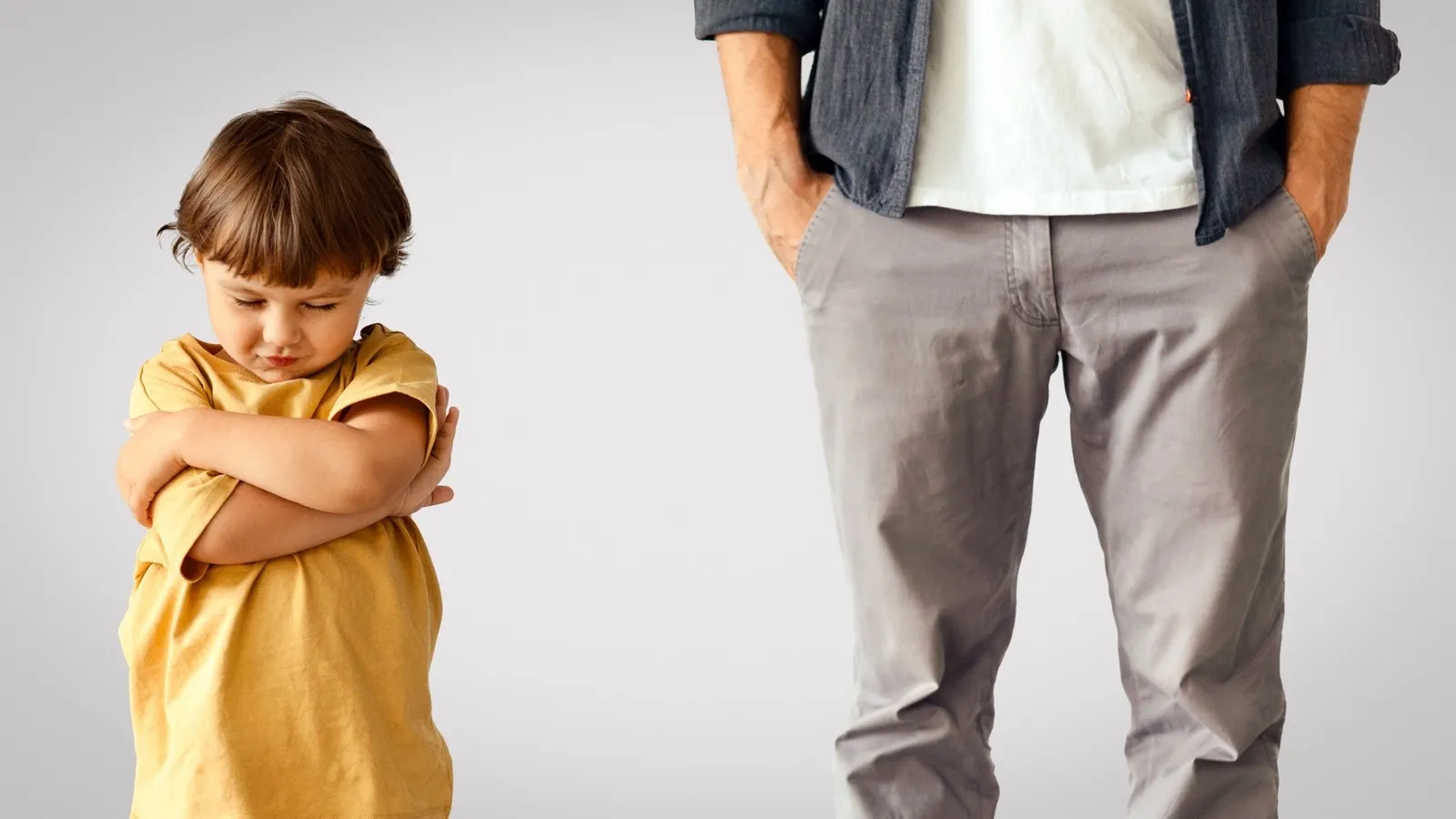 Offended little boy standing with crossed hands, unrecognizable indifferent man standing behind with