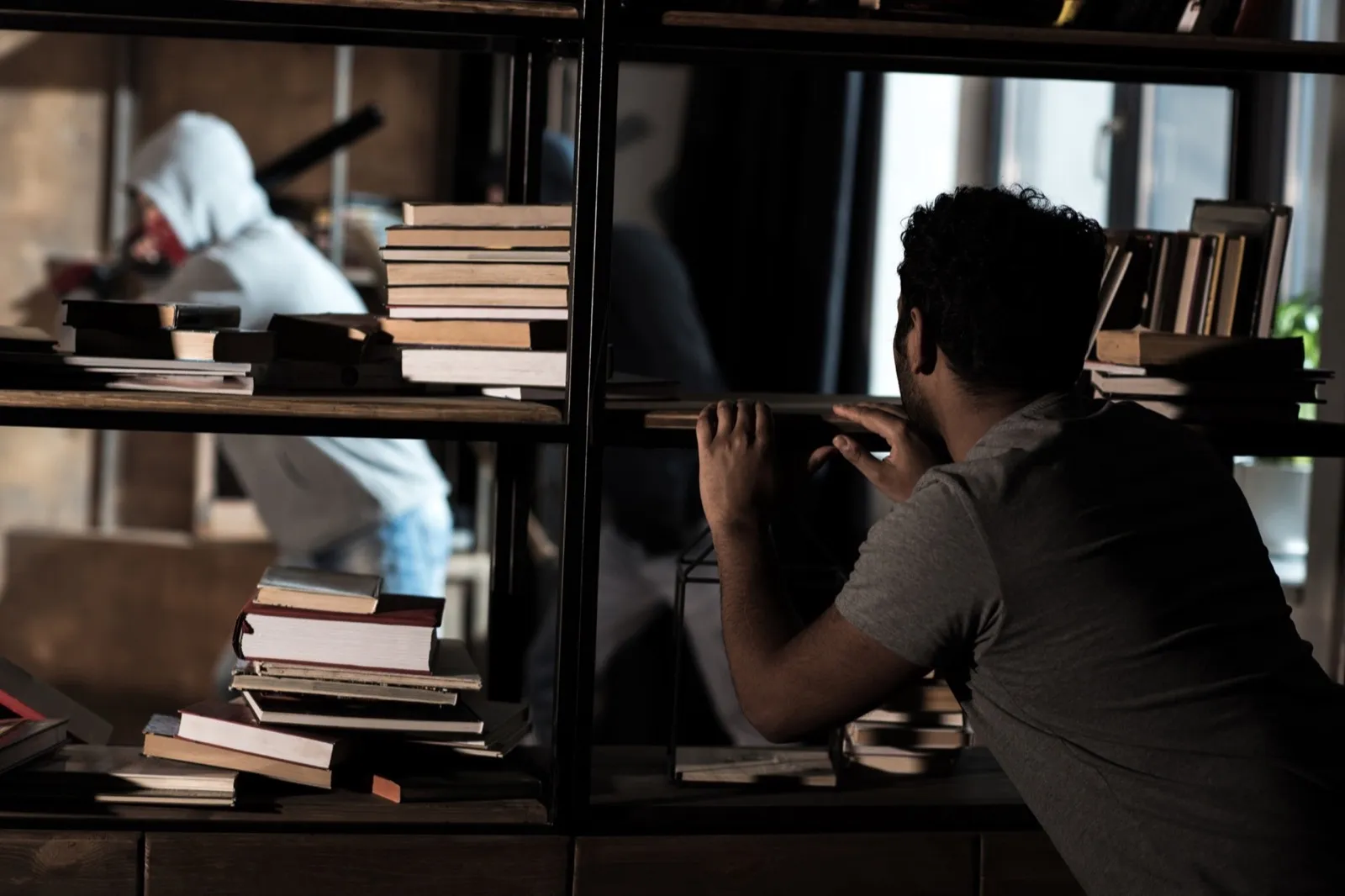 Scared man hiding from burglars behind bookshelves, house robbery scene