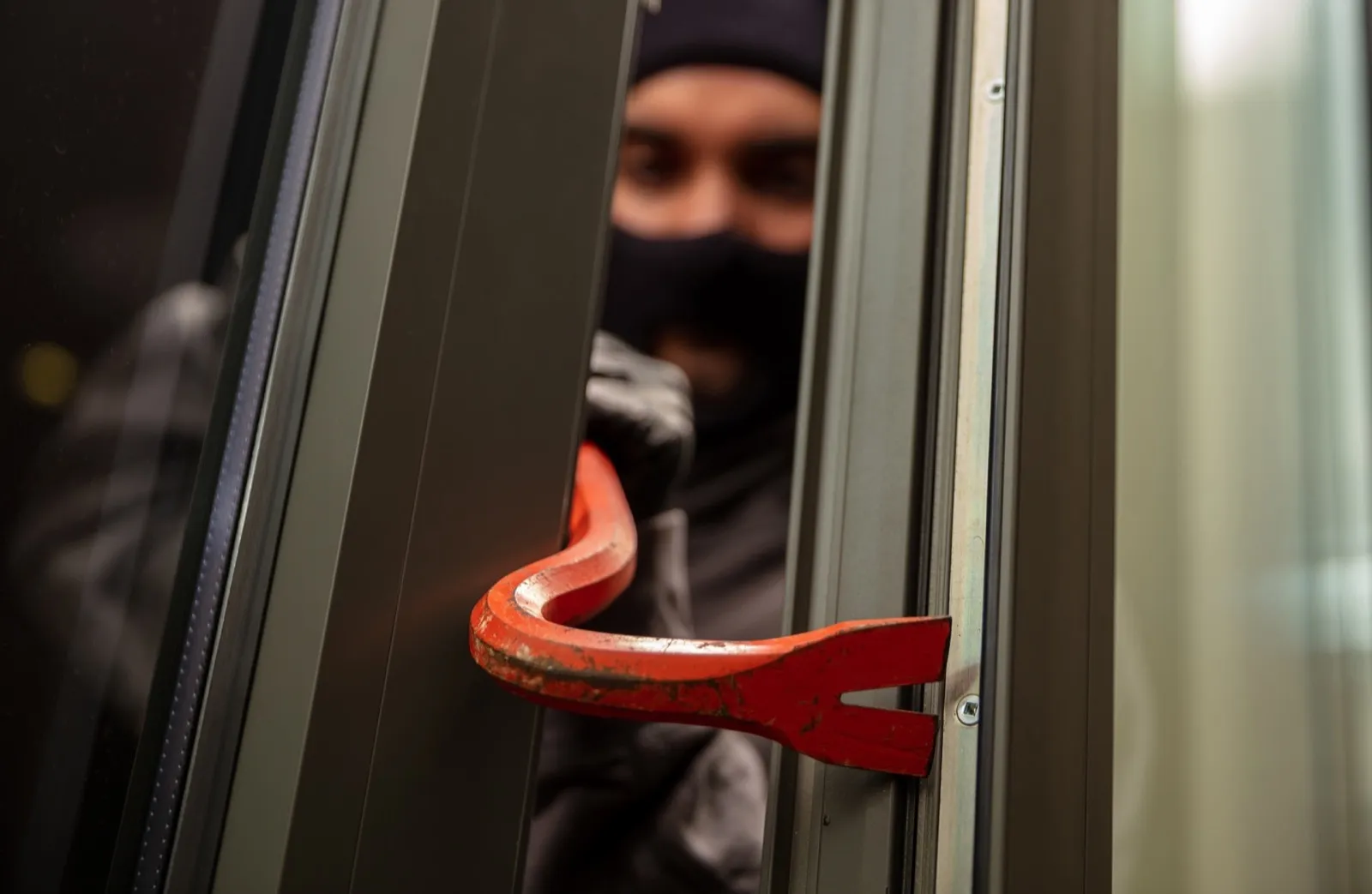 Thief in balaclava holding a crowbar to break a glass window.