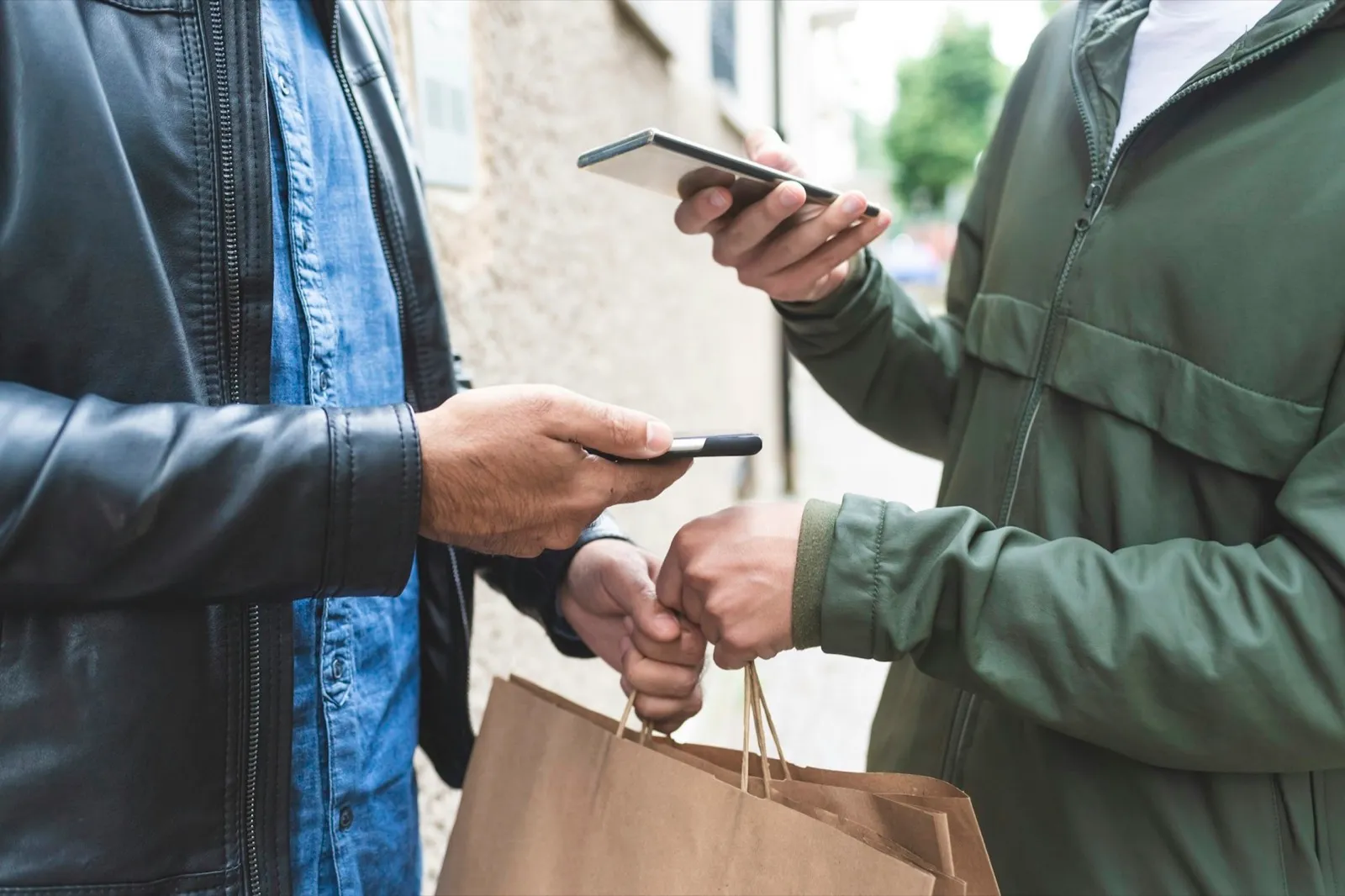 Two people exchanging mobile phones and a paper bag on the street.