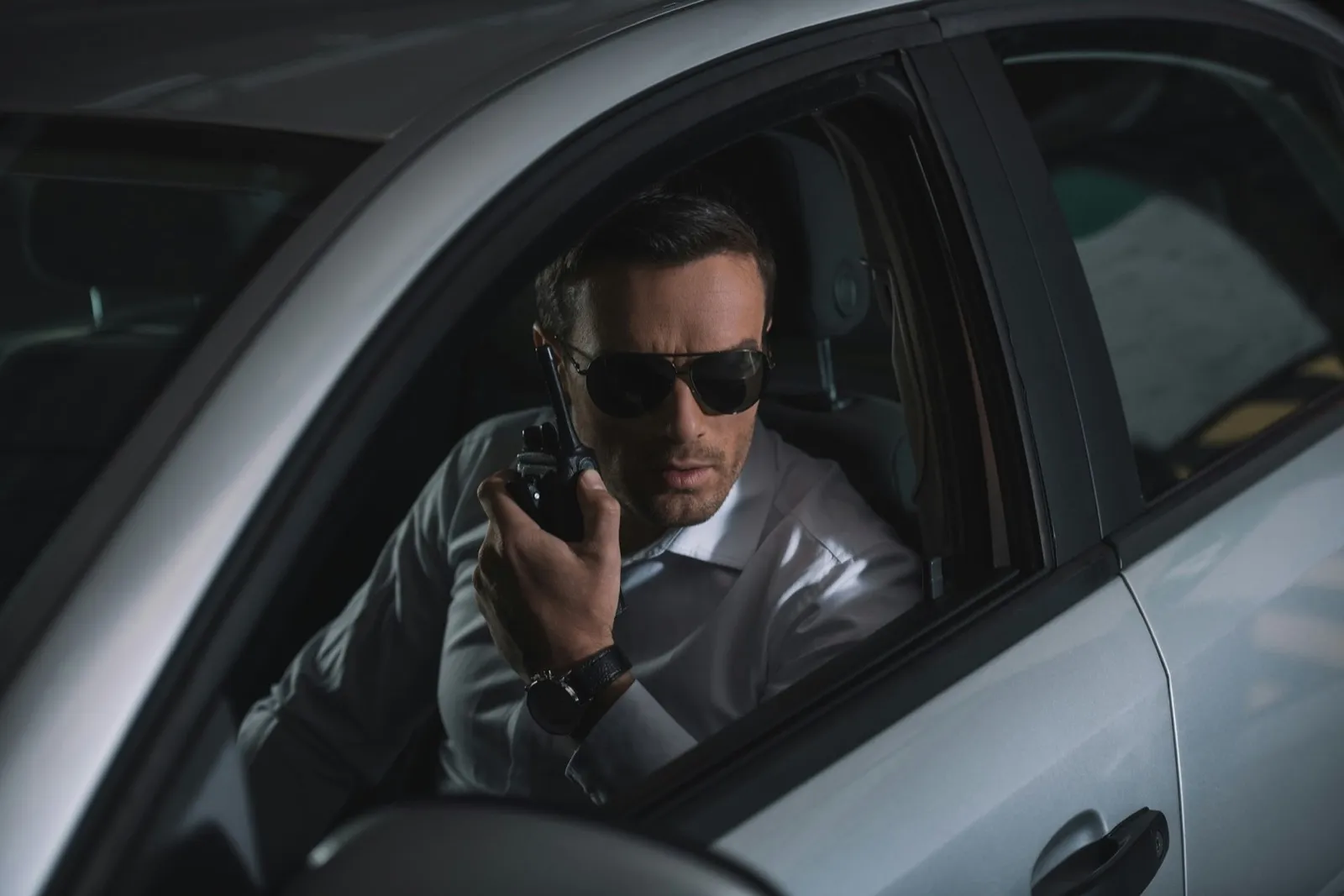 undercover male agent in sunglasses doing surveillance and using talkie walkie in car
